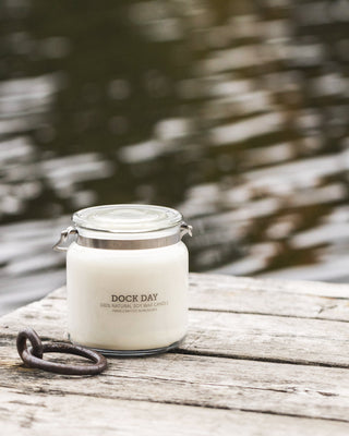 Dock Day candle on a weathered wooden dock with a metal dock mooring ring, lake water in soft focus behind.