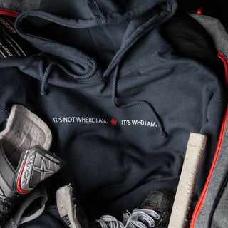 Black It's Not Where I Am. It's Who I Am. hoodie surrounded by hockey equipment.